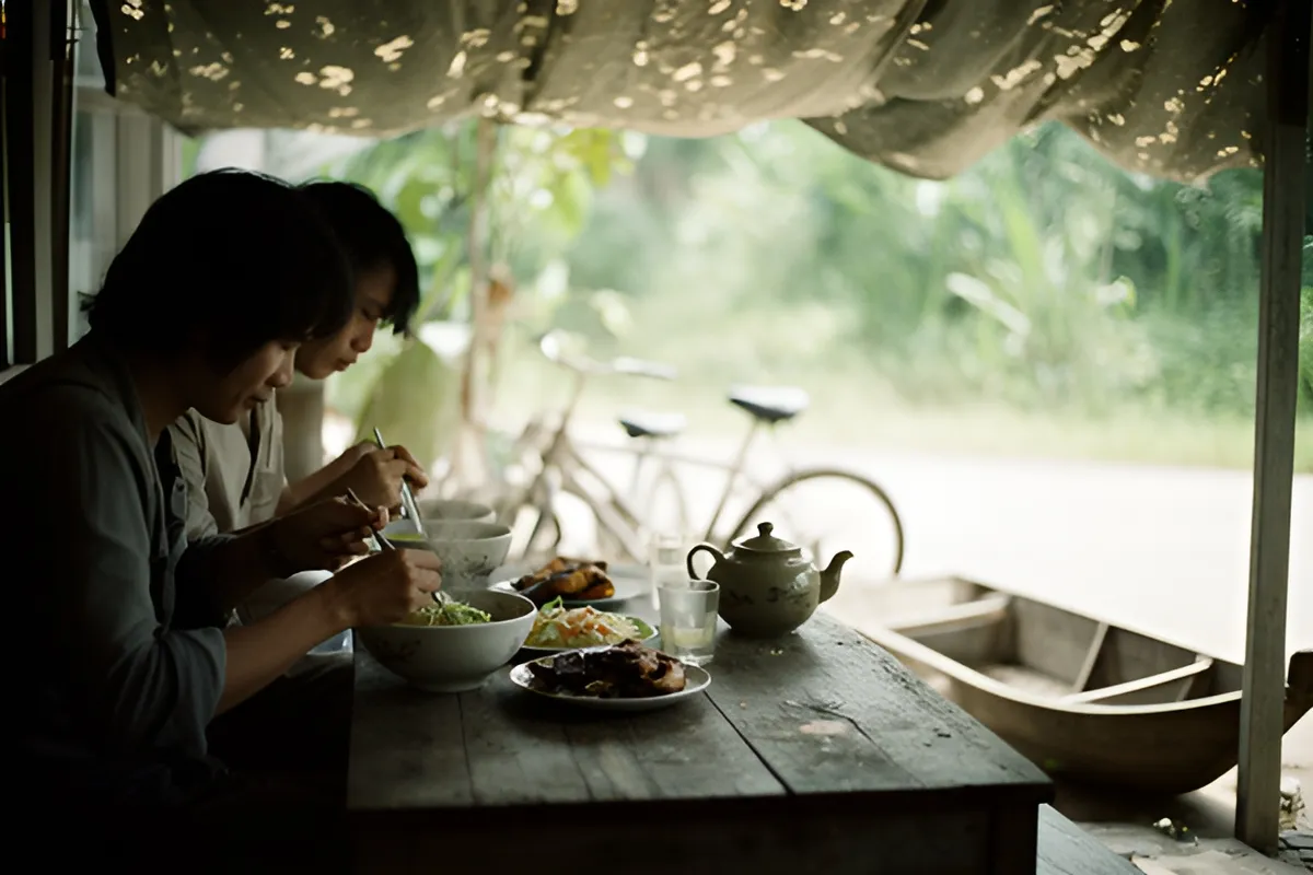Hai người ngồi ăn mì bên bàn gỗ dưới mái che, trên bàn có ấm trà, ly và đĩa thức ăn, phía sau có xe đạp và thuyền nhỏ