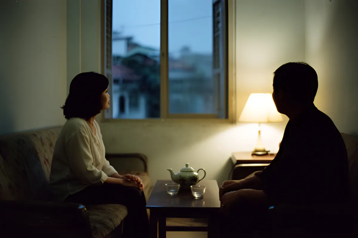 Hai người ngồi im lặng trên ghế sofa trong phòng khách buổi tối, bàn thấp có ấm trà và hai chén, đèn bàn sáng vàng bên cửa sổ