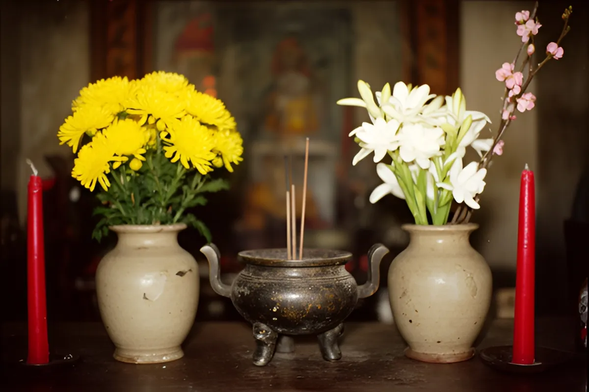 Bàn thờ với hai bình gốm cắm hoa vàng và hoa trắng kèm cành hoa hồng, giữa là bát hương đồng, hai ngọn nến đỏ tắt