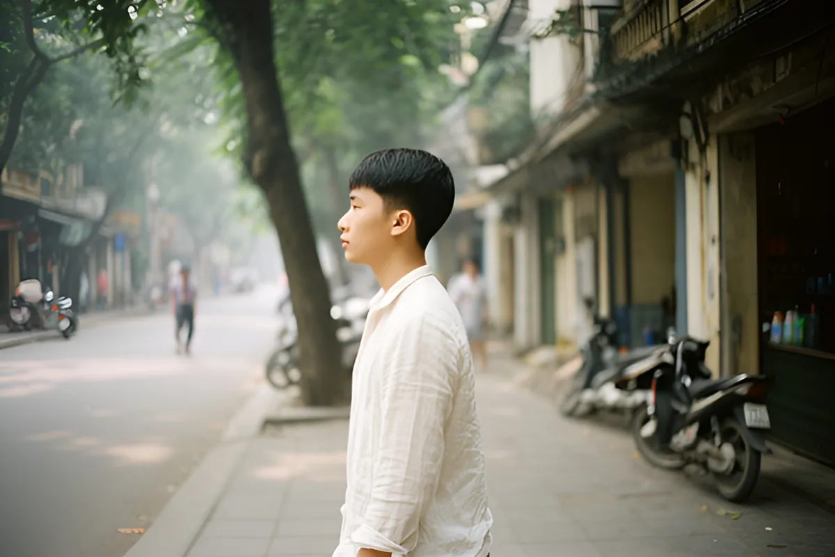 Một thanh niên mặc áo sơ mi trắng đi bộ trên vỉa hè phố có cây xanh, xe máy đậu ven đường, vài người ở xa