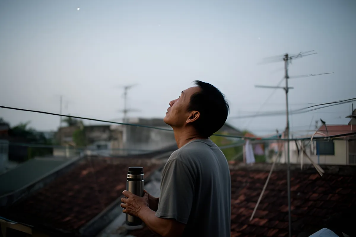 Một người đàn ông đứng trên mái nhà lúc sáng sớm, ngước nhìn bầu trời, hai tay ôm một bình giữ nhiệt