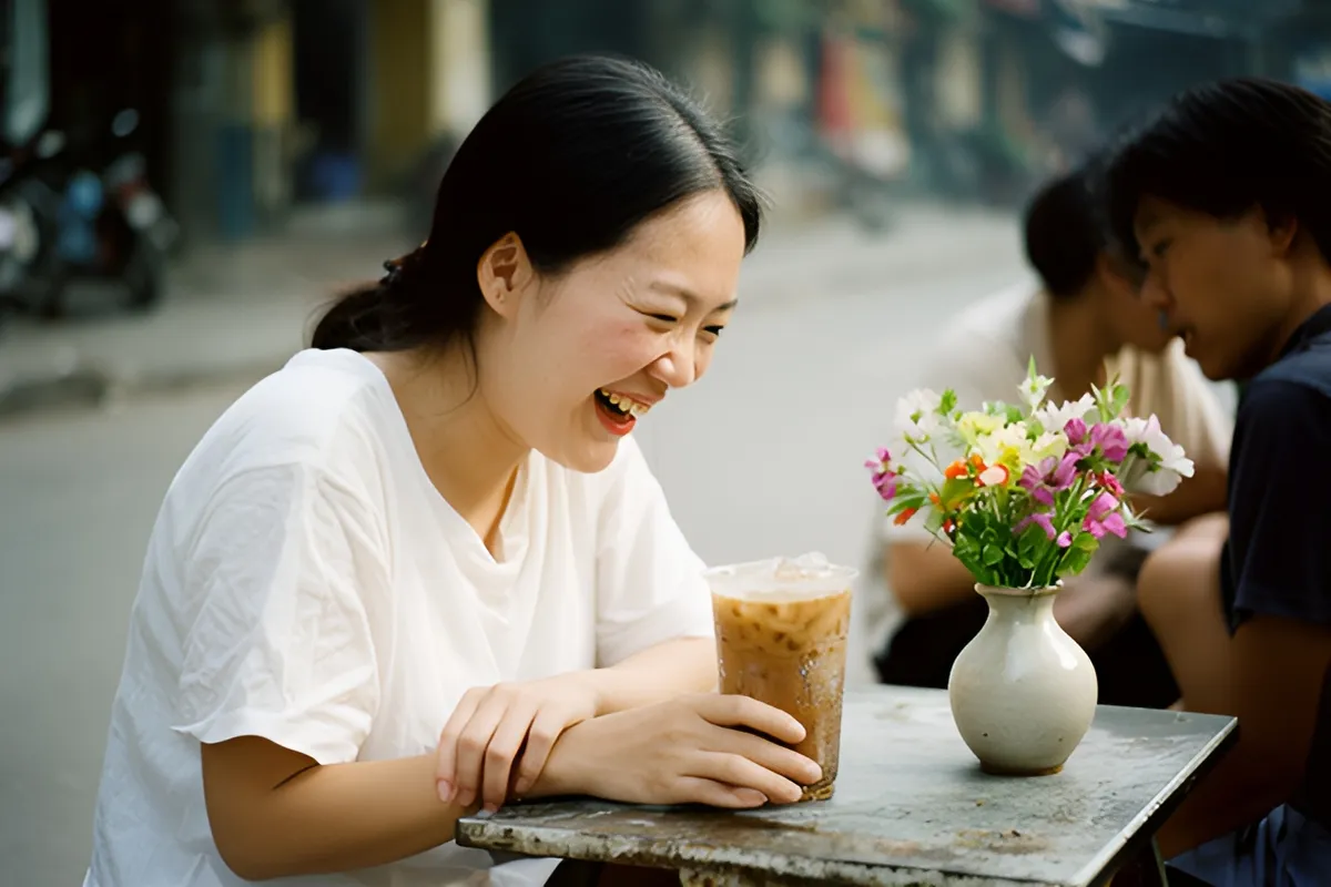 Một phụ nữ trẻ áo trắng cười tươi nhìn xuống ly cà phê sữa đá trên bàn kim loại nhỏ, cạnh đó là bình hoa nhỏ