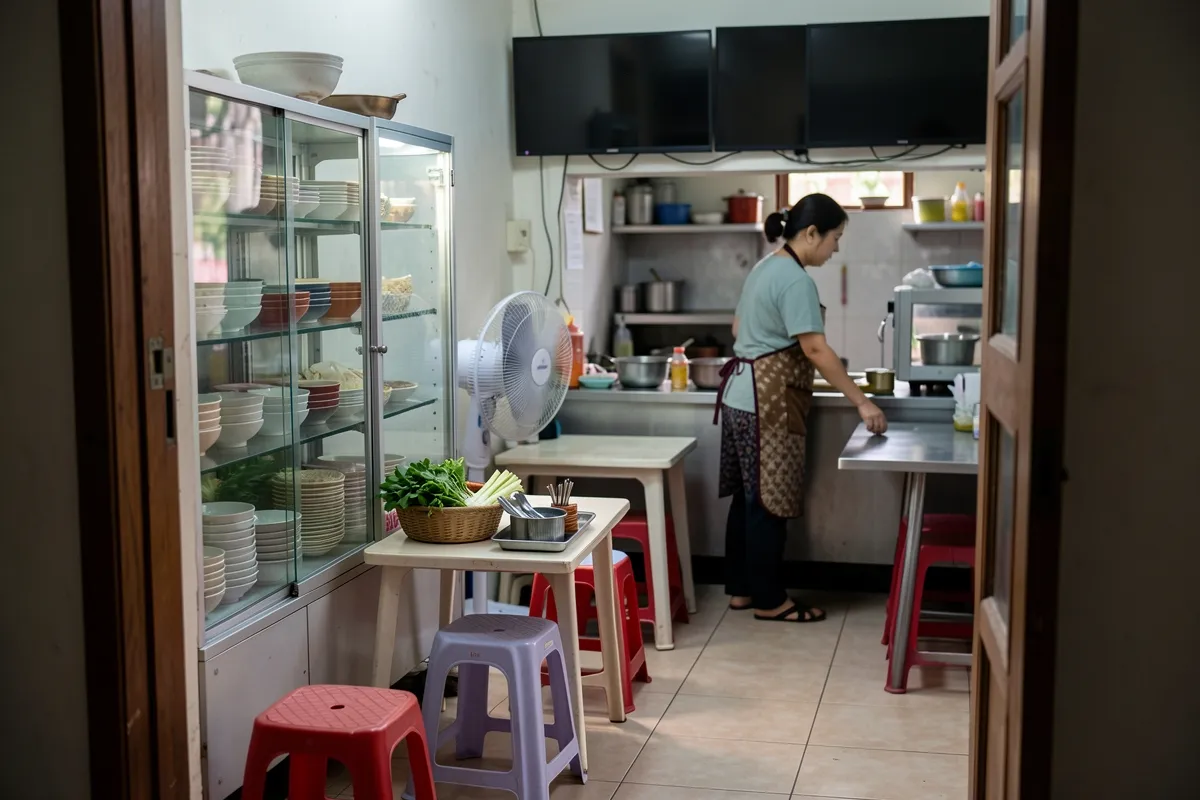 Một phụ nữ mặc tạp dề đứng lau bàn trong gian bếp nhỏ, bên cạnh tủ kính chứa bát đĩa, quạt đứng và ghế nhựa