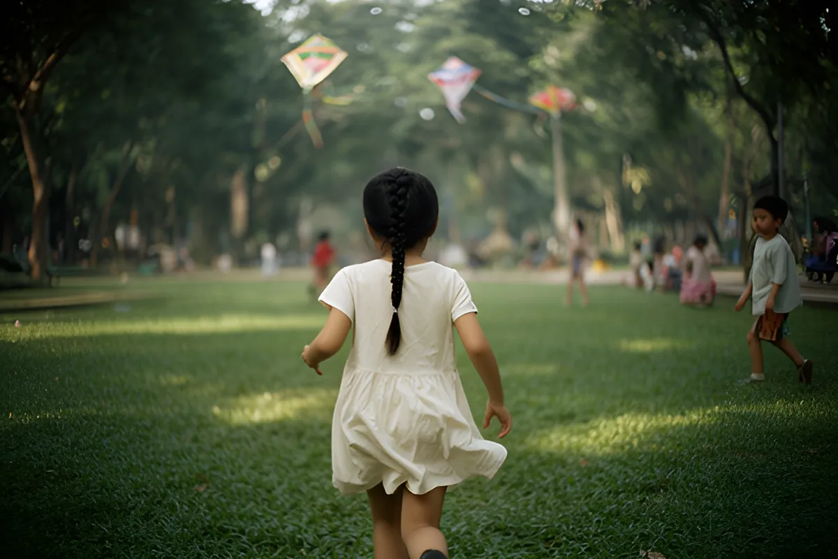 Một bé gái tóc đuôi bện chạy trên bãi cỏ công viên, bên cạnh có một bé trai đi bộ