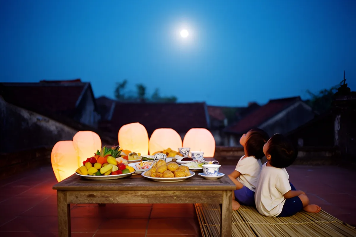 Hai em nhỏ ngồi trên chiếu ngoài sân nhà mái ngói, ngước nhìn trăng sáng, bên cạnh bàn gỗ thấp bày trái cây