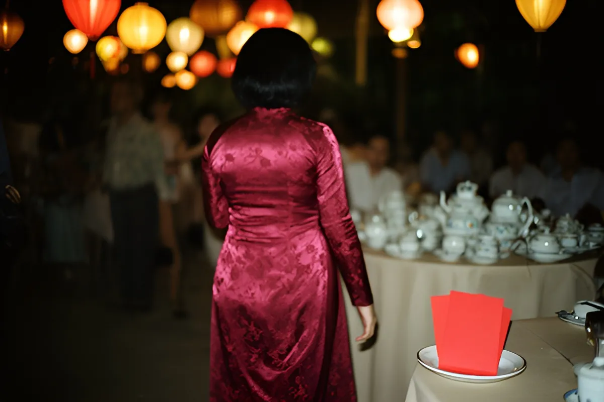 Người phụ nữ tóc ngắn mặc áo dài đỏ thêu hoa quay lưng, sau lưng là đèn lồng và bàn trà trắng, trước có đĩa phong bì đỏ