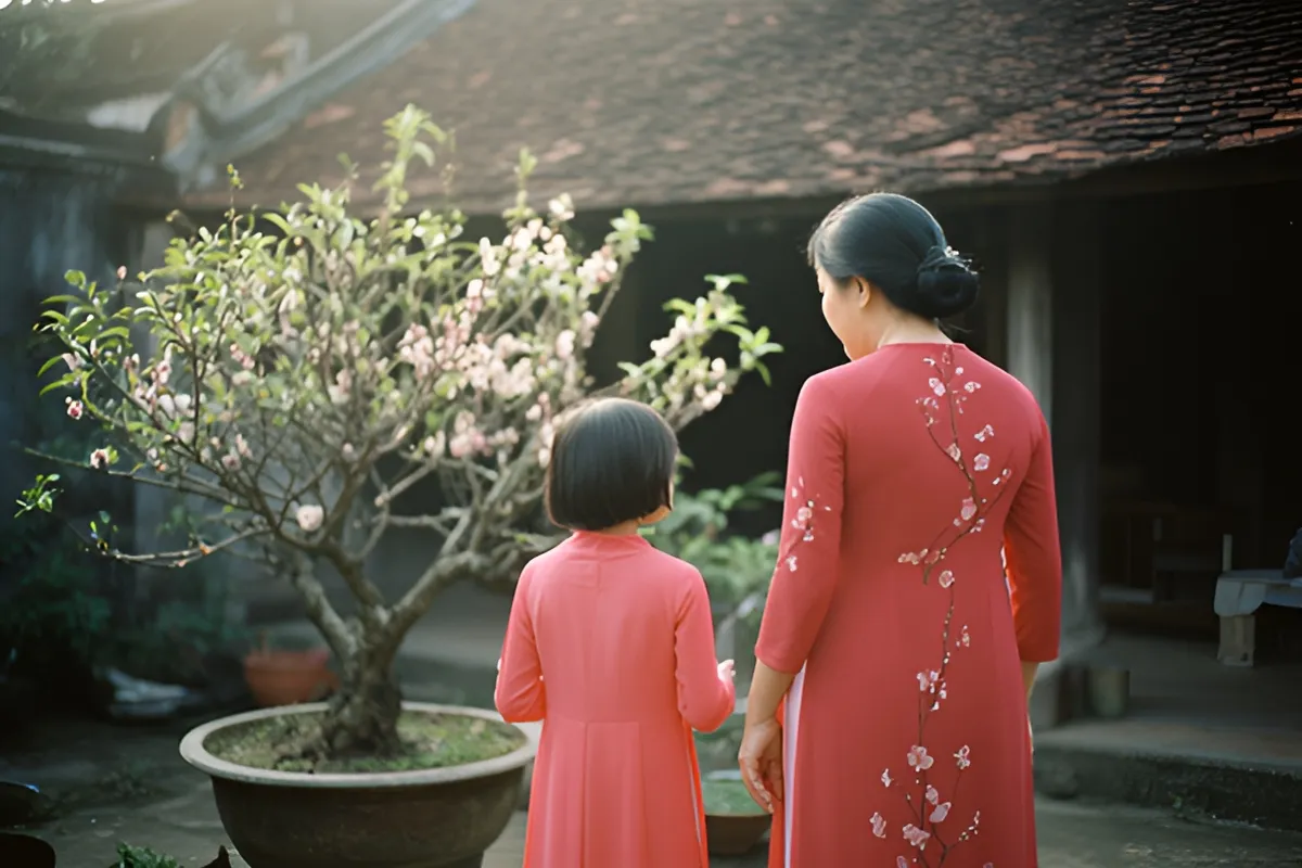 Hai người phụ nữ mặc áo dài đỏ đứng quay lưng nhìn cây hoa trong chậu ở sân nhà ngói, ánh sáng chiều dịu nhẹ