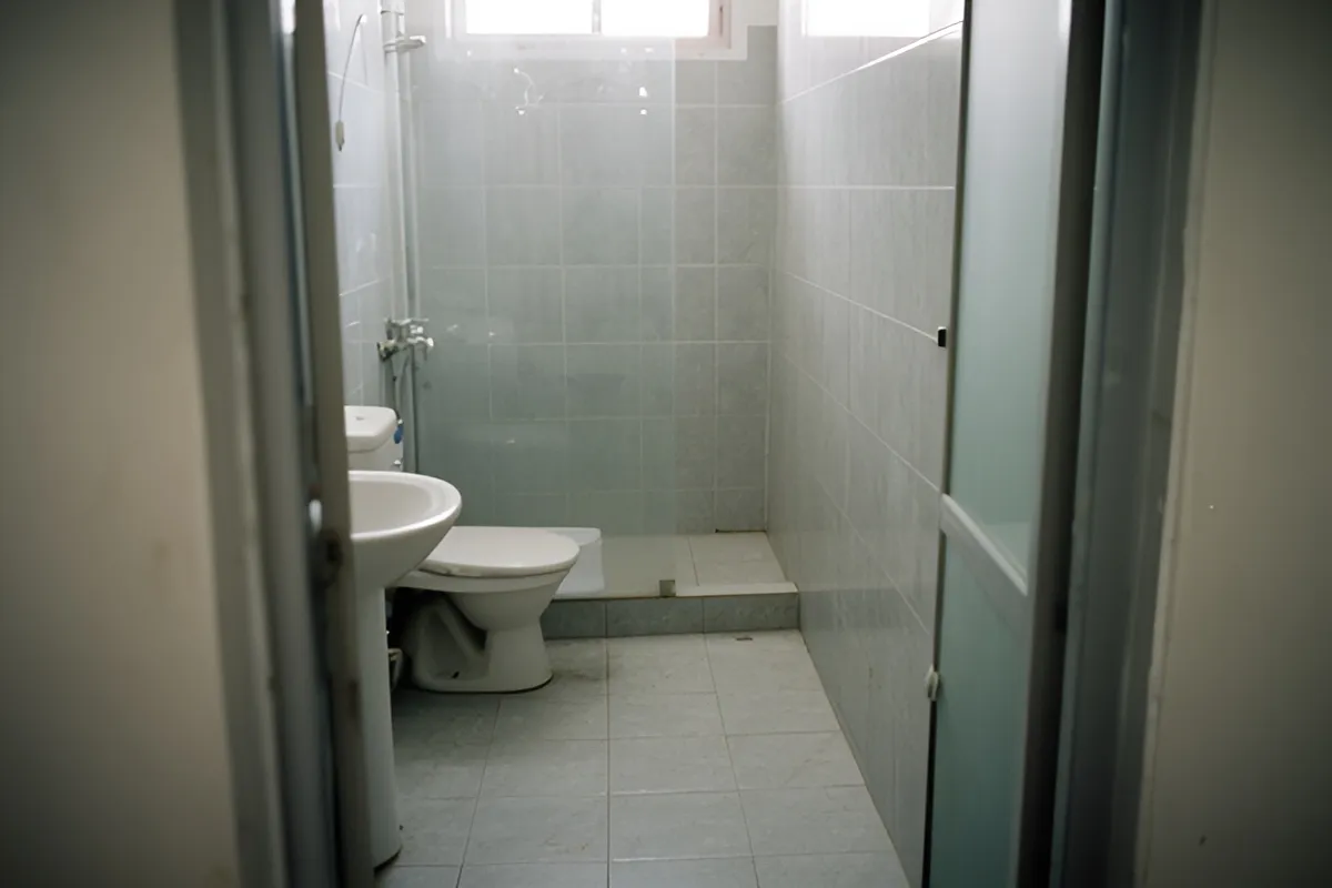 Phòng vệ sinh nhỏ lát gạch xám, có bồn rửa, bồn cầu và khu tắm đứng ngăn bằng kính, ánh sáng dịu từ cửa sổ