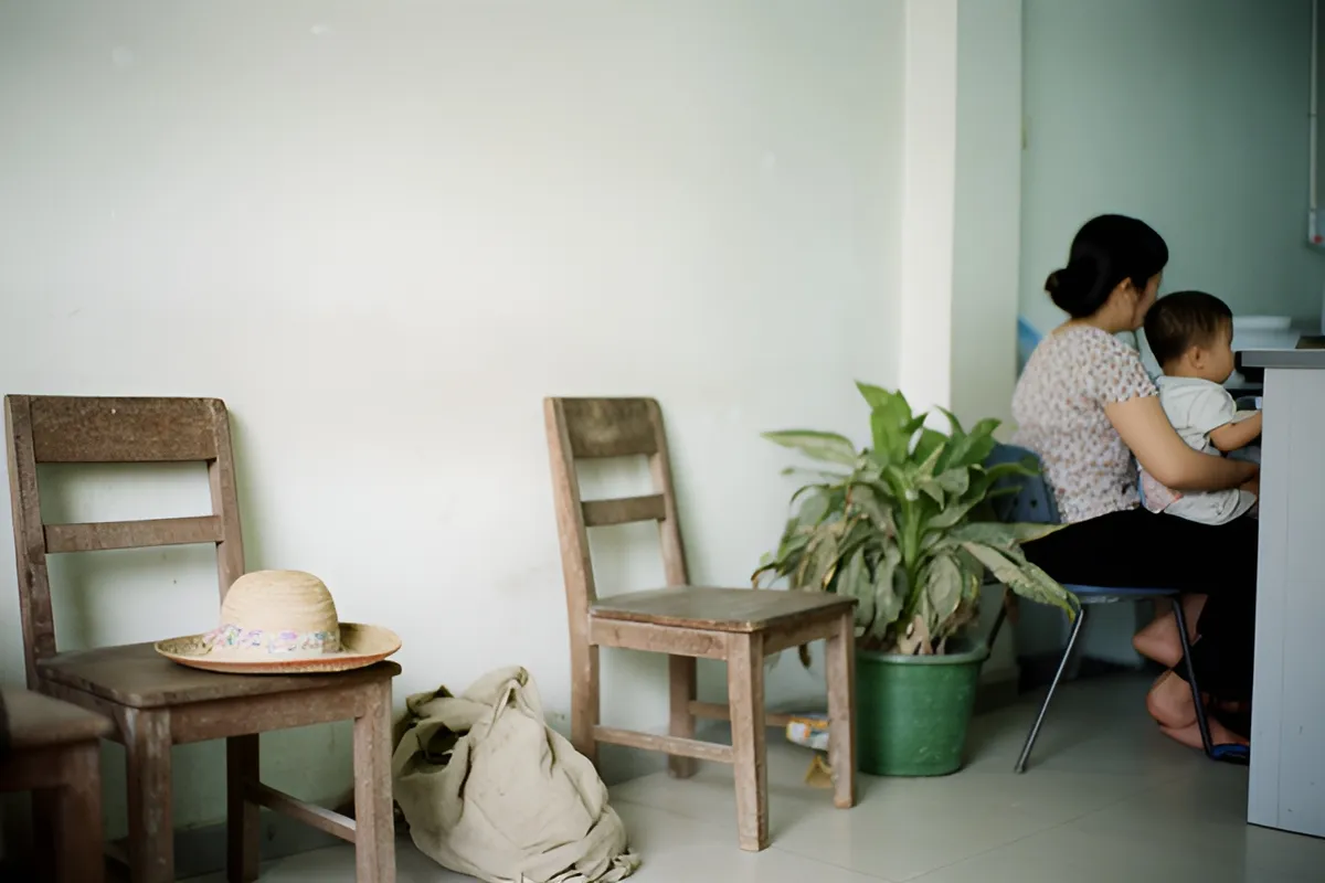 Một người phụ nữ ngồi ôm đứa nhỏ trên đùi ở góc phòng sáng nhẹ, cạnh hai ghế gỗ cũ