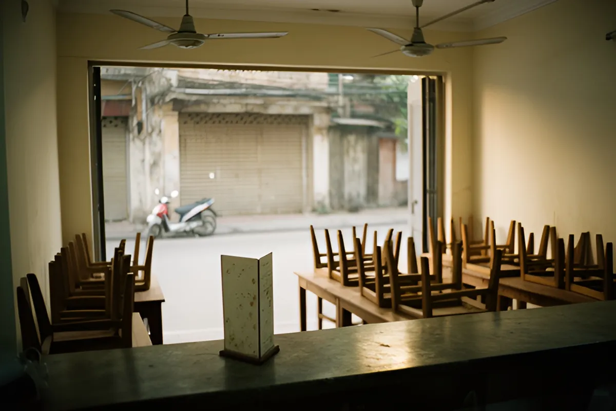 Quán ăn vắng người buổi chiều, ghế gỗ úp trên bàn, quạt trần tĩnh lặng, ngoài phố có xe máy đỗ và cửa cuốn đóng