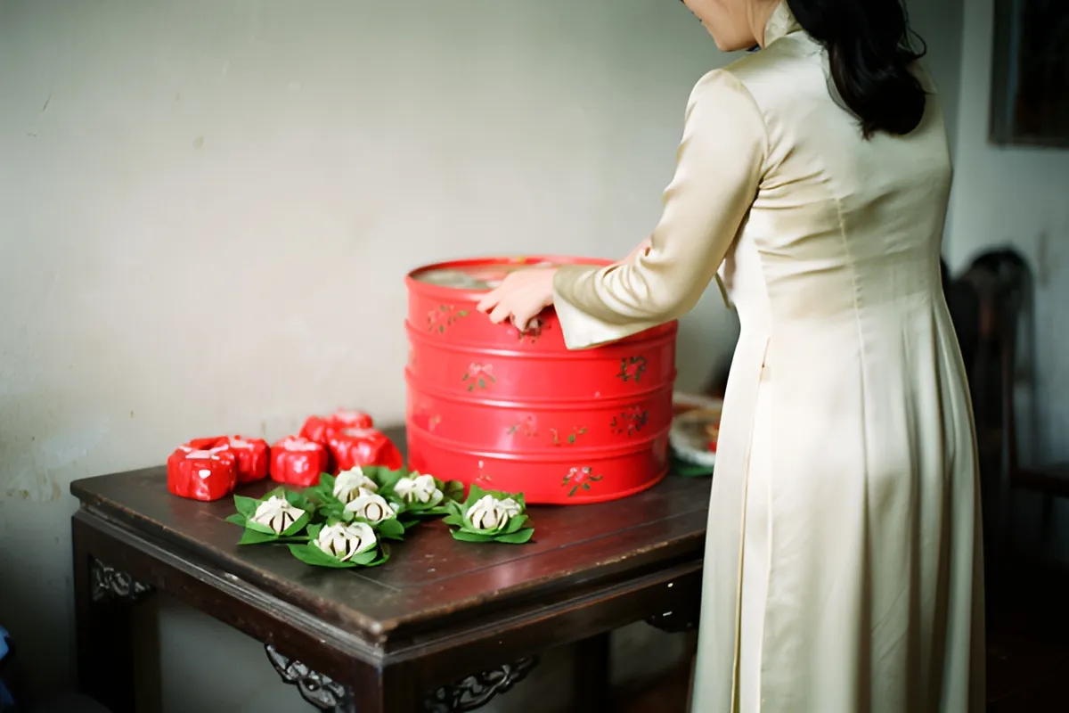 Một người phụ nữ mặc áo dài màu kem đứng cạnh bàn gỗ, tay đặt lên chồng khay tròn đỏ có hoa văn