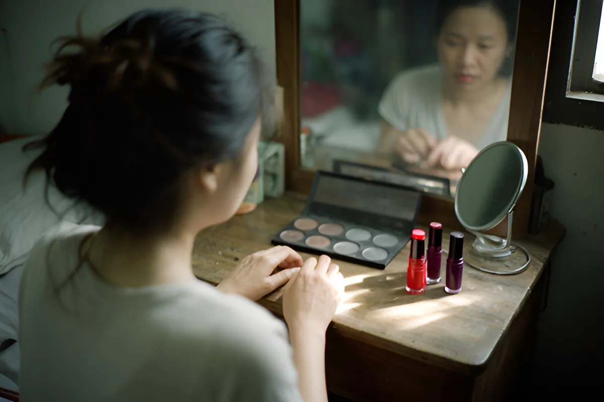 Một người phụ nữ tóc búi ngồi quay lưng trước bàn gỗ có gương, tay đặt trên mặt bàn cạnh hộp phấn mắt và vài chai sơn móng