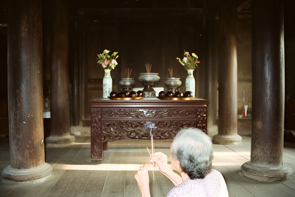 Một bà cụ tóc bạc quay lưng đứng thắp nhang trước bàn thờ gỗ chạm khắc giữa nhà sàn gỗ với cột gỗ dày