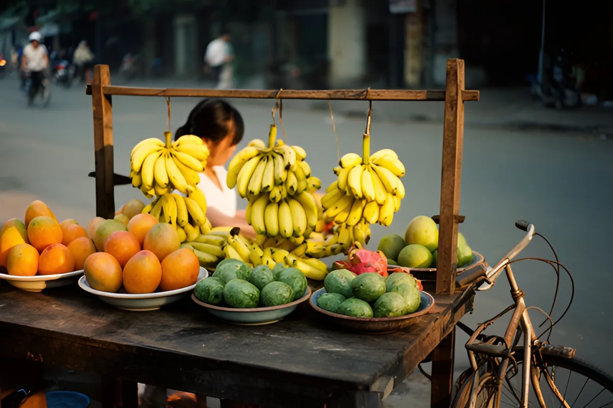Một phụ nữ đứng sau quầy gỗ bán trái cây với các chùm chuối vàng treo trên khung, khay xoài và quả xanh, xe đạp cũ ở mép phải