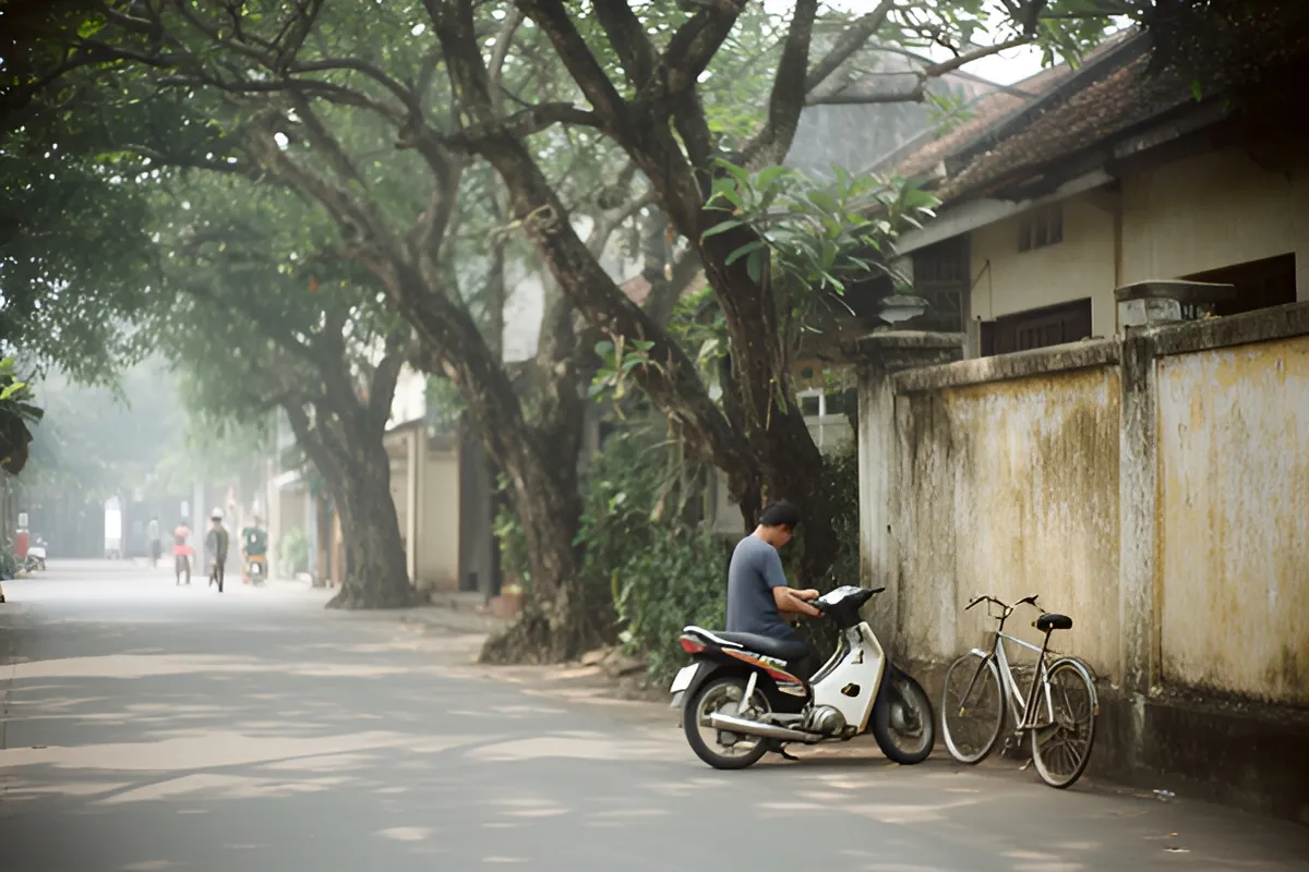 Một người đàn ông ngồi trên xe máy đậu ven tường, cạnh một chiếc xe đạp