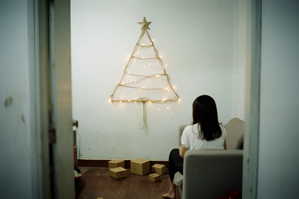 Một người phụ nữ tóc đen ngồi trên ghế sofa nhìn lên cây thông dây đèn treo tường, dưới sàn có vài hộp các-tông nhỏ