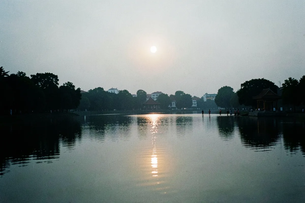 Vài người đi bộ từ xa bên bờ hồ nước phẳng lặng, mái đình và bóng cây in trên mặt nước, mặt trời thấp chiếu vệt sáng dài