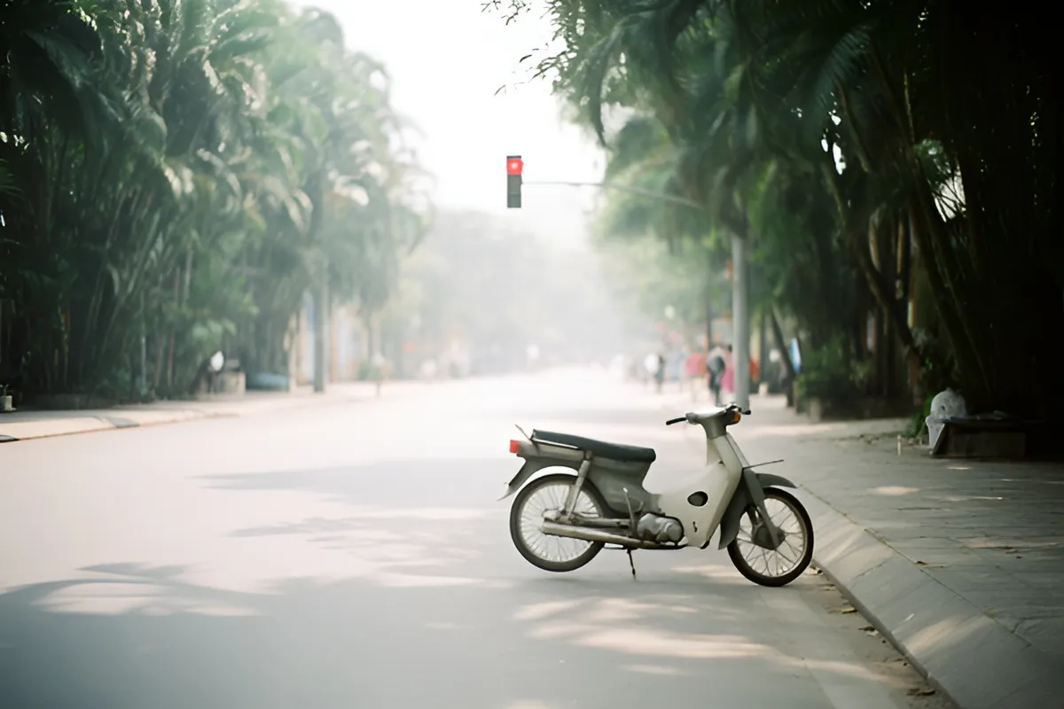 Một chiếc xe máy trắng dựng bên vỉa hè, đường dài hai bên là hàng cây, xa xa có đèn giao thông đỏ, ánh sáng ban ngày