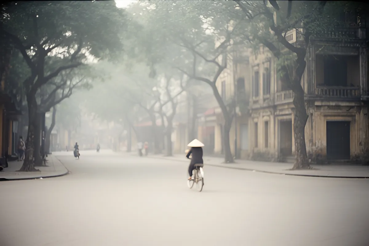 Một người đội nón lá đạp xe giữa đường vắng, vài người ở xa, hai bên là cây cổ thụ và nhà cổ, sáng sớm sương mờ
